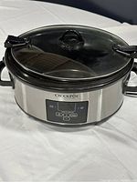 Front view of Crock-Pot The Original slow cooker showing stainless steel body, digital control panel, and clear glass lid with black latch handles.