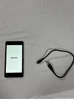 Front view of Sony Xperia M4 Aqua smartphone powered on showing Sony logo, lying next to black charging cable with micro USB connector.