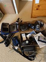 Photo showing assortment of cameras including Minolta SRT 101 with Tamron lens, Canon Elph, Pentax digital camera with damaged card cover, Yashica film camera, Optex video light, GPX portable CD player, cords, cases, and manuals