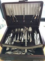Community Plate flatware set in a brown canteen, containing forks, knives, spoons, and serving pieces organized in the case.