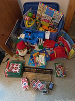 Wide view of the lot showing toys in blue plastic bins, books, card games, cribbage board, and holiday décor arranged on the floor.