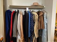 Full view of many coats and clothing items hanging on a rack including fur coat, leather coat, patterned coats, polka dot shirt, and hat displayed on top shelf.