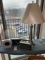 Photo showing 30-inch tall metal lamp with beige shade, Panasonic RF-2400D radio, Sylvania radio, and white sunrise alarm clock on table near window.