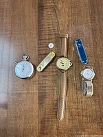 Overall view of the lot showing stopwatch, 2 wristwatches, 2 pen cutting tools and button battery on wood surface.