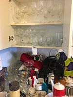 Photo inside cupboard showing stacked clear stemware wine glasses, some short and some tall, various clear drinking glasses on lower shelf, mugs, and a black Keurig coffee maker on counter.