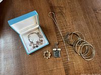 Full lot displayed on wooden surface including silver charm bracelet, four bangles, etched bracelet, pendant with chain, and seed pearl pin.