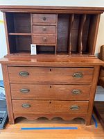 Front view of the antique wooden desk cabinet showing upper small drawers, open shelves, vertical file holders on the right, three lower drawers with metal oval handles, and overall wood finish with scratches and wear.