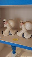 Two white ceramic sailor figurine savings banks with large sacks on their backs shown together on shelf.