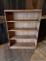 Front view of wooden four-shelf bookcase on wooden floor with barn wall background, showing wood grain and open shelves.