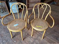 Two vintage bentwood chairs with cane woven seats placed on wooden floor with some scratches visible on wood surfaces. Chairs have looped backs and curved armrests.