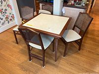 Photo of square folding wooden card table with cream vinyl insert, surrounded by four folding cane back chairs with cushioned seats on hardwood floor.