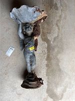 Cast metal angel cherub bird bath basin lying flat on a concrete surface, showing the figure holding the basin above its head with dirt accumulation.