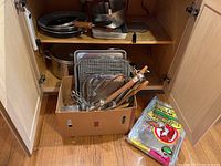 Cookware items stored in cabinet including frying pans, metal pots, and a box with grilling utensils and baking trays.