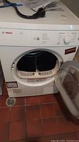Front view of white Bosch Axxis dryer with door open and power cord resting on top, tiled floor beneath