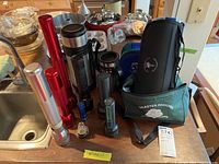 Photo showing all items in lot including flashlights of various sizes, travel cups, carafes, lunch bags, and travel blanket on kitchen counter near sink.