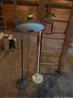Two metal base floor lamps side by side on a wooden floor in a barn. One black with cone-shaped shade and flexible arm, the other white with dish-like shade and straight pole. Visible rust and wear.