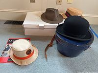 Overview of four hats and three hat boxes arranged on floor. Visible are black Dobbs bowler hat atop blue Dobbs box, brown fedora, straw hat with red/black band, and cream bucket hat with orange band and 1915 logo.