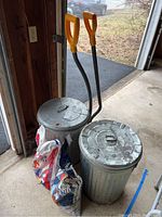 Two galvanized metal trash cans with lids and two yellow-handled snow shovels next to them, with bags of rock salt and ice melt on the floor.