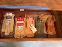 Seven pairs of leather gloves in a drawer, some packaged, some unpackaged, varied colors from beige to tan and green, showing all gloves included in lot.