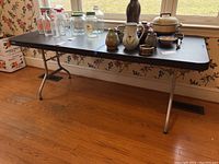 Full view of the Lifetime folding table with multiple glass and ceramic items on top for scale, showing black textured plastic top and metal legs.