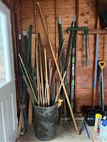 Full view of the metal can filled with various garden stakes made of metal, wood, and bamboo leaning inside a shed against a wooden wall.