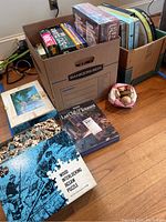 Photo showing two banker’s boxes filled with boxed puzzles of different sizes and types, along with a partially assembled wood interlocking jigsaw puzzle and a sealed puzzle box lying on the floor.