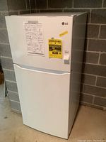 Front and side view of white LG refrigerator with top freezer and bottom fresh food compartment on basement floor.
