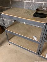 Full view of metal shelving unit with particle board shelves positioned against concrete block wall, showing overall structure and condition.