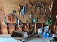 Overall view of pegboard with assorted yard hand tools, coils of wire, two bags of Kingsford charcoal, two blue bottles of windshield washer fluid, and rusted metal wheelbarrow on floor beneath pegboard.