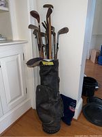 Full view of the vintage black leather golf bag standing upright filled with 15 wooden and metal shaft golf clubs.