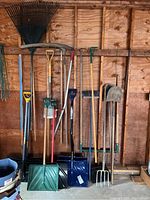 Full view of various garden and yard tools including rakes, shovels, snow shovels, fork, broom, and a picker hanging on a wooden wall in a garage.