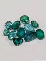 Loose genuine emerald and blue gemstones of varying shapes and sizes laid on white background.
