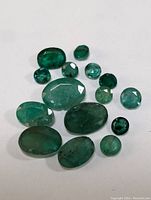 Fourteen loose emerald gemstones of various sizes and shapes laid out on a white surface.