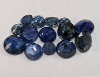 Close-up image showing fourteen loose blue sapphire gemstones of varying shapes including oval, round, pear and cushion cuts, different sizes, and color depths ranging from medium to dark blue.