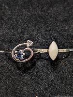 Top view showing the two silver rings on black background; one with blue gemstone and clear smaller stone, the other with white marquise-shaped stone.