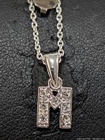 Close-up photo showing the 'M' shaped pendant with clear cubic zirconia stones embedded on its surface, hung on a sterling silver chain with link design.