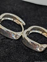 Close-up photo of a pair of polished sterling silver hoop earrings with one clear round stone each, positioned on black background.