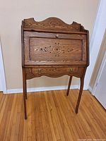 Full view of antique wooden secretary desk in closed position showing carved design on drop front and decorative top panel.