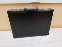 Closed black leather briefcase resting on a white surface, showing front and side view with handle and combination locks.