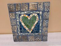 Full frontal view of square mosaic piece with central green heart and patterned tile border on beige wall background.