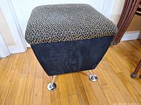 Photo of the foot stool showing the cushioned top with leopard print fabric and black upholstered sides resting on chrome legs.