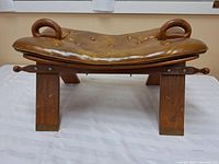 Side view of the leather foot stool showing leather seat, brass feet on wooden legs, and curved wooden handles.
