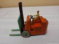 Side view of the orange metal Dinky toy forklift with green forks and seated driver figure on white background.
