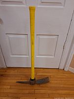 Full view of the pick axe with yellow handle against white door, showing length and condition