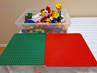 Photo showing clear bin filled with assorted vintage Lego Duplo large building blocks and toy parts placed on red and green base plates.