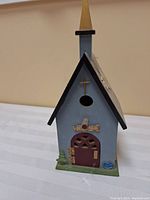 Full view of wooden bird house showing blue body, black roof edges, circular bird entrance hole, wooden spire on top, and decorative details including mini tree, welcome sign and blue bird figure on green base.