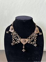 Front view of the vintage gilt brass necklace displayed on a black bust, showing the intricate embossed triangular drops connected by double chains with hanging bombé-shaped charms.