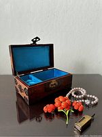 Open wooden Asian jewelry box showing interior lining and one side compartment, accompanied by coral flower brooches, faux pearl necklace, and metal key.