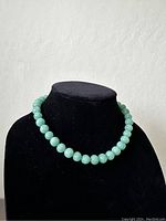 Front view of a necklace composed of smooth, round green beads displayed on a black bust stand.