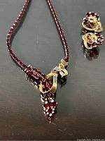 Red stone necklace with matching clip-on earrings laid out on a dark surface showing the decorative bow on the necklace and the earrings' red stones and gold clasp.
